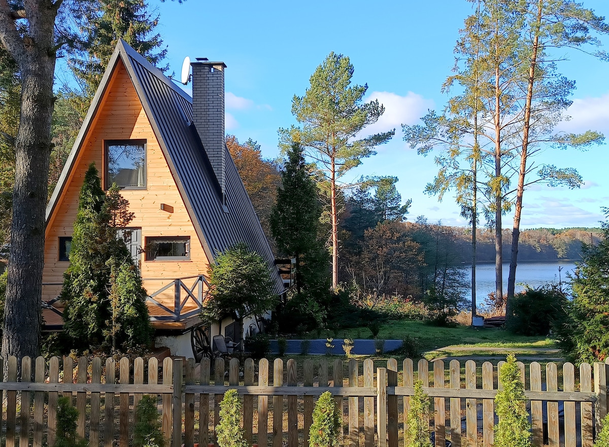 The exterior of a triangular wooden cabin is set against a backdrop of trees and a serene lake. A wooden fence encloses the property, with lush greenery surrounding the house. The sloped roof features a chimney, complementing the peaceful natural surroundings.