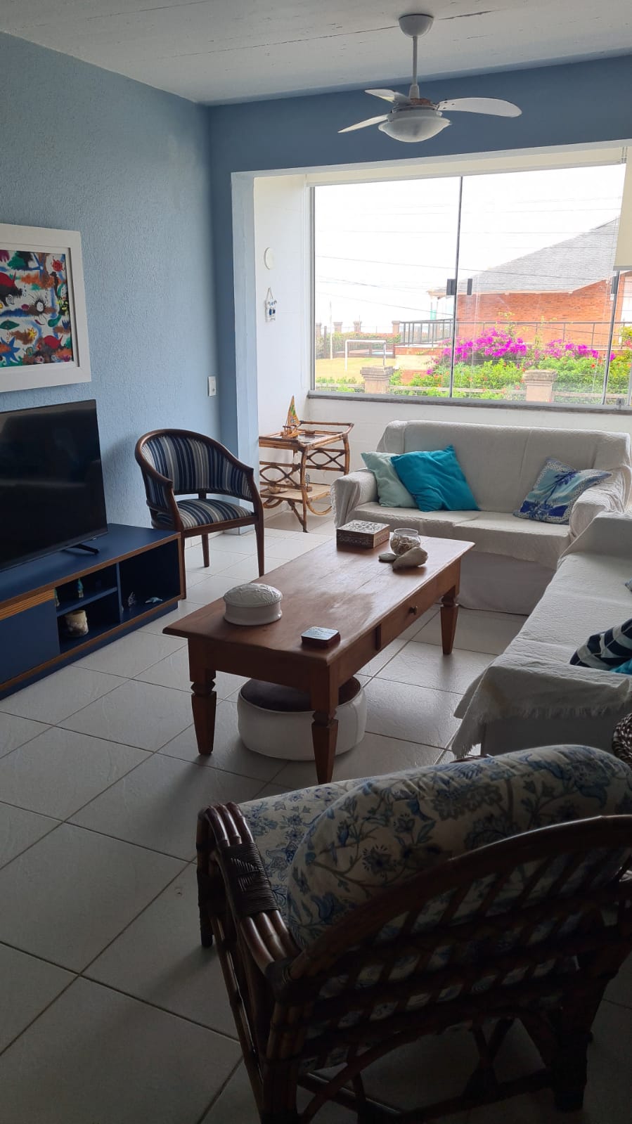 A cozy living area features a light-colored sofa and wicker chairs, complemented by a wooden coffee table. A Smart TV is positioned against the wall, while a vibrant piece of wall art adds a touch of color. Large windows provide natural light and views of the outdoor area.