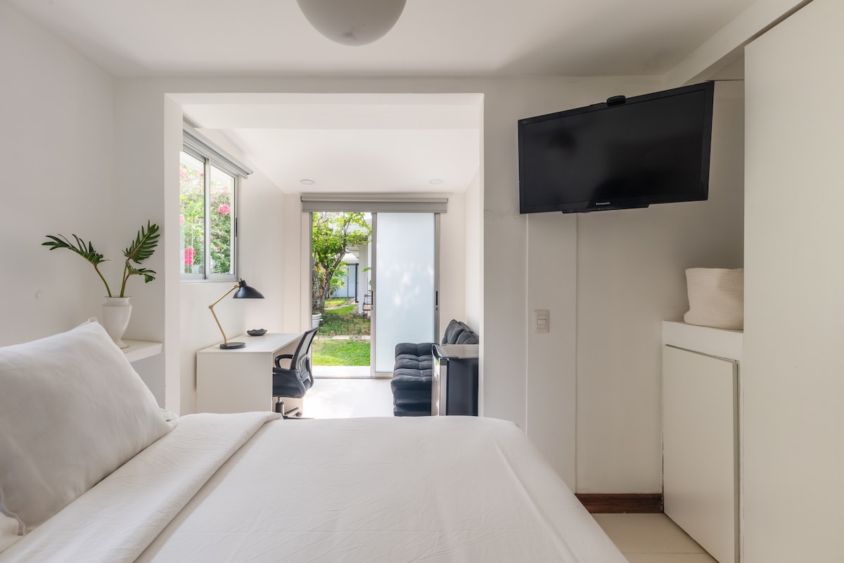A bright room is presented, featuring a spacious bed with crisp white linens. A Smart TV is mounted on the wall opposite the bed. Light flows in through large windows that lead to a view of a green garden and a workspace with a desk and chair.