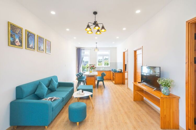 A modern living area features a teal sofa and a small round table, accompanied by a TV mounted on a wooden stand. Four dining chairs surround a kitchen table in the background, with natural light illuminating the space from windows adorned with light curtains.