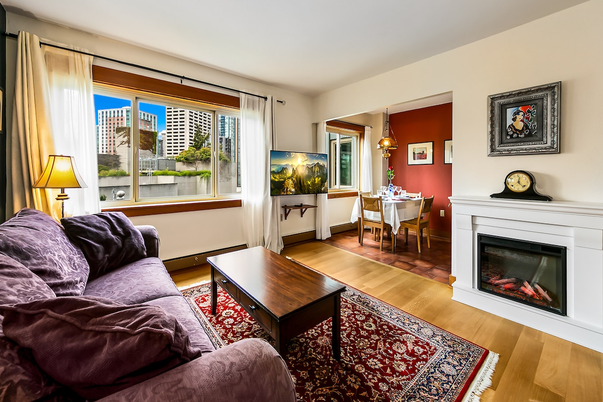 A cozy living area features a plush purple couch and a wooden coffee table, illuminated by natural light from large windows. An electric fireplace adds warmth, while a dining space with a table for four is seen through an archway. A vibrant rug enhances the inviting atmosphere.