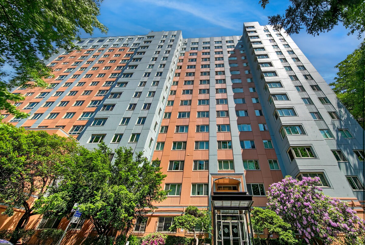 A multi-story condominium building is depicted, featuring a combination of orange and gray exterior panels. Lush greenery surrounds the base of the building, complemented by blooming purple flowers. Large windows reflect natural light, contributing to the building's modern appearance.