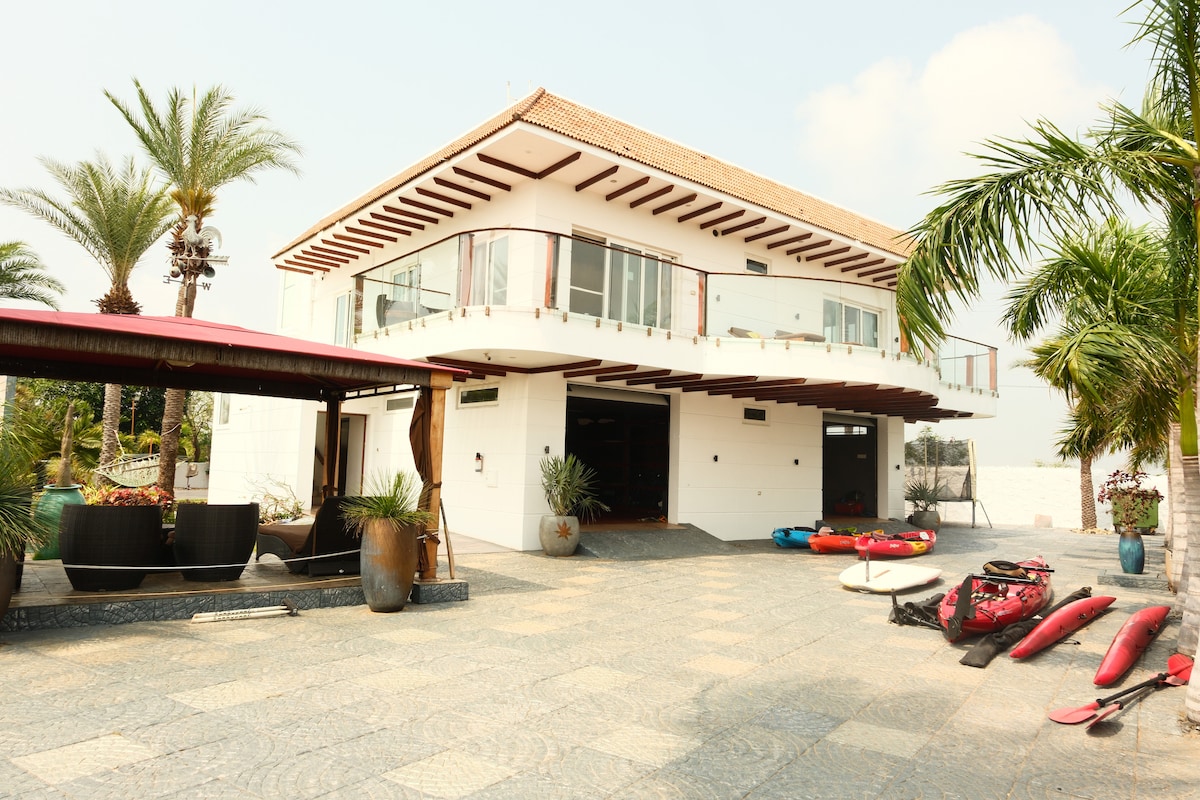 The modern villa features a two-story structure with large glass windows and a spacious outdoor area. Palm trees and potted plants surround the patio, where colorful kayaks are neatly arranged. The design incorporates a covered seating area with a vibrant awning, enhancing the inviting atmosphere.