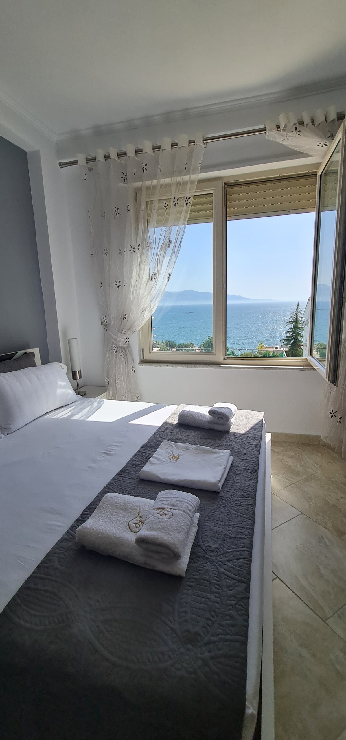 An inviting bedroom features large windows that showcase a sea view. Soft linens are arranged neatly on a spacious bed, complemented by rolled towels placed on top. Light drapes filter the sunlight, creating a bright and airy atmosphere within the room.