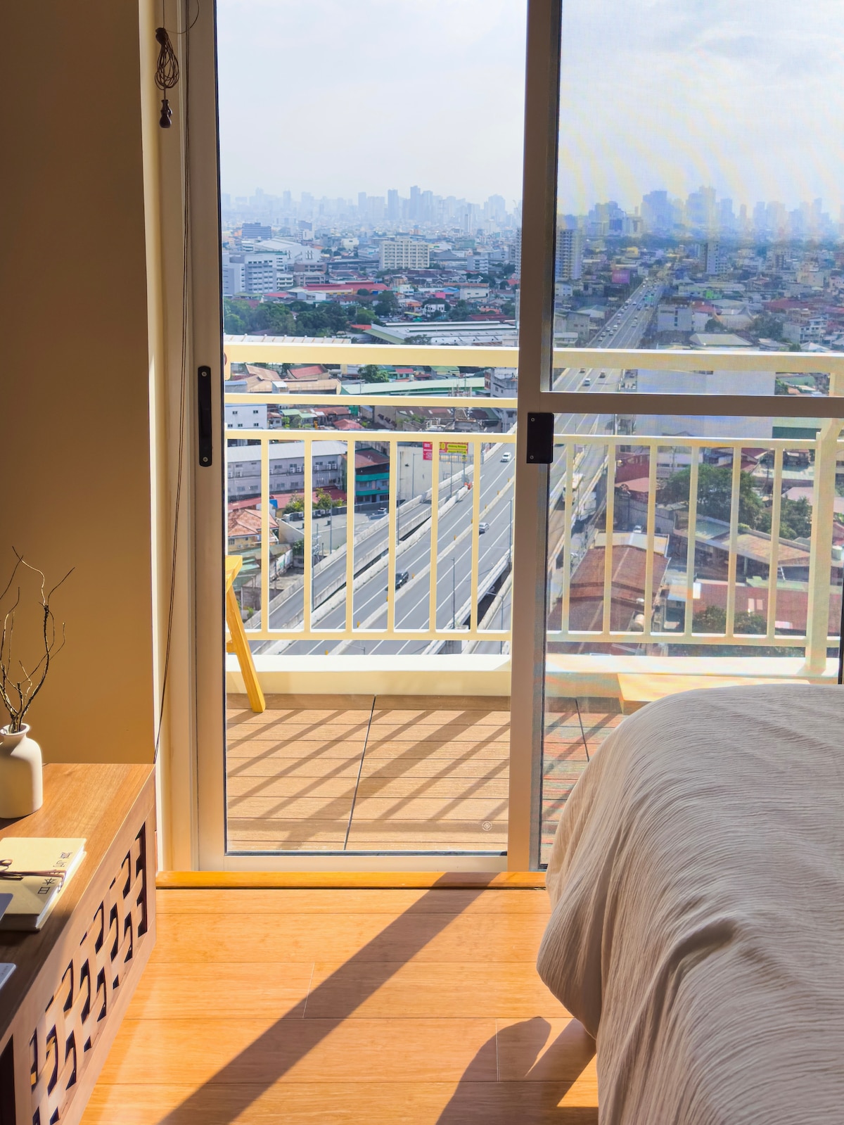 A balcony is visible with a railing framing a city skyline in the distance. Light enters through sliding glass doors, illuminating a wooden bedside table and a spacious bed. The soft colors of the interior create a serene atmosphere.