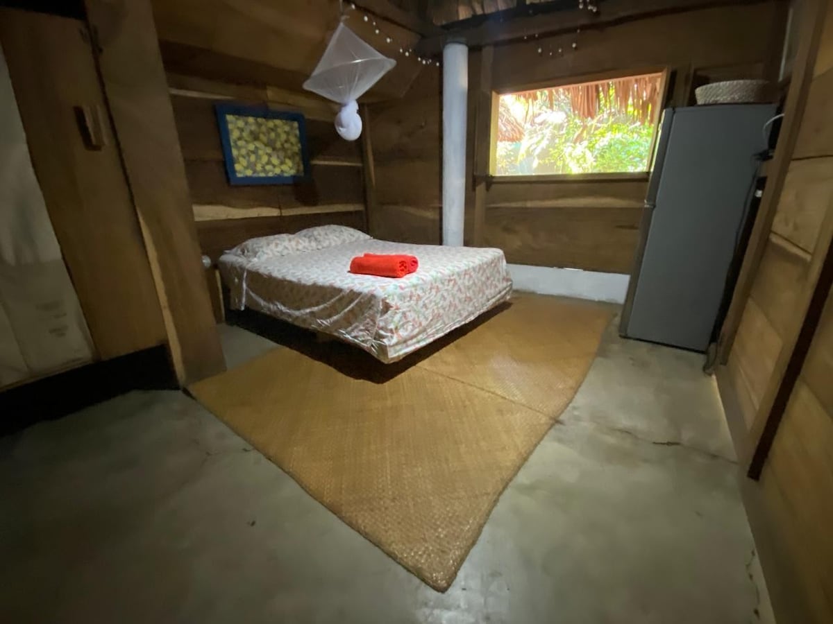 The room  is equipped with a mosquito net, fridge/freezer, floor fan and looks out onto a private terrace and tropical garden. There is an attached on-suite bathroom with toilet and sink.