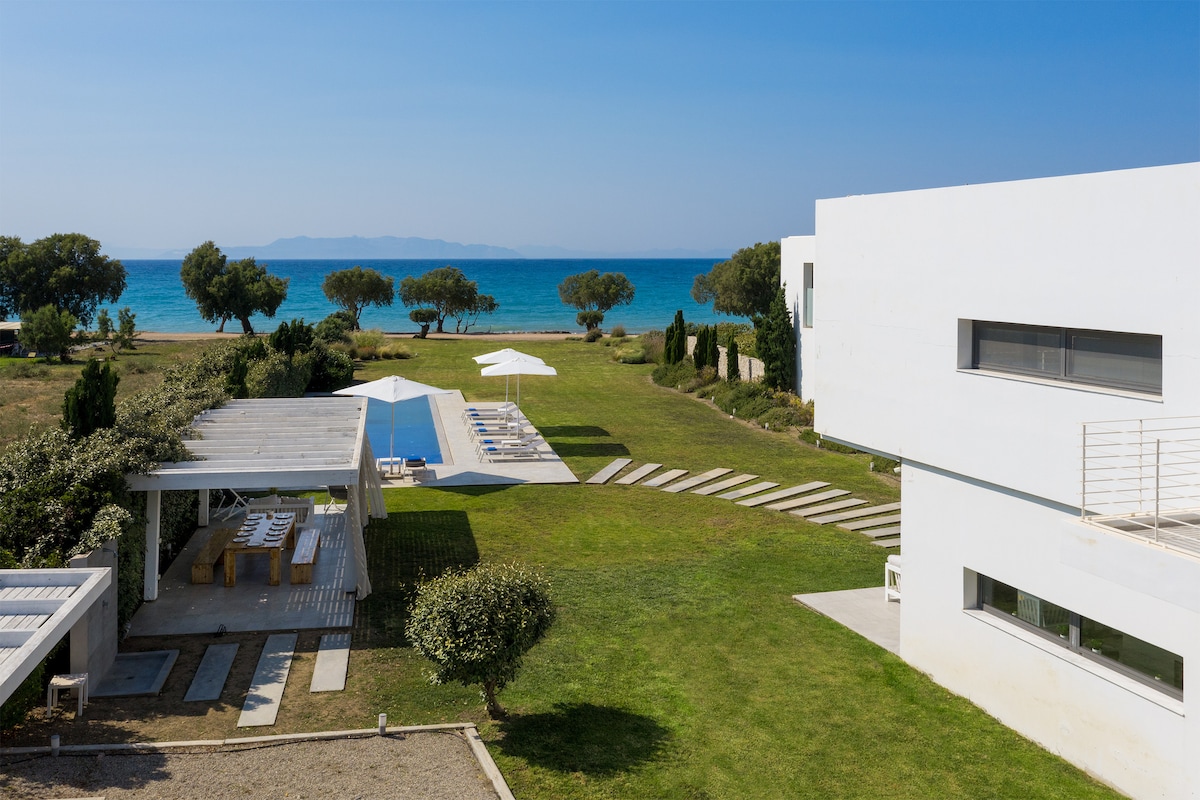 A wide view of the serene outdoor area highlights a well-kept lawn leading to a mosaic pool and a beachfront. Sun loungers are arranged near the pool, while a shaded dining pergola is positioned nearby. The clean lines of the modern villa are visible in the background.