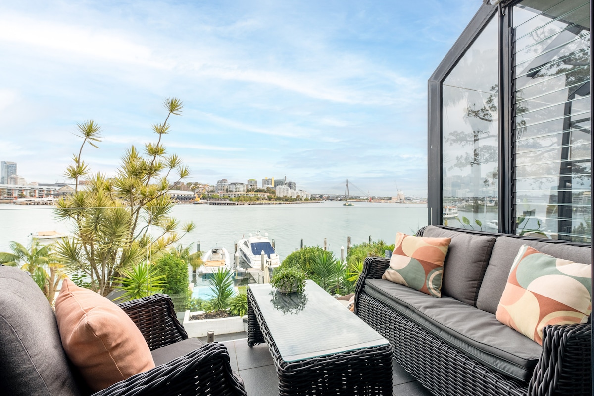 A waterfront balcony features comfortable seating on a modern wicker sofa, accented with colorful cushions. A glass coffee table is positioned in front, with views of boats and lush greenery across the river. Clear skies enhance the serene atmosphere.