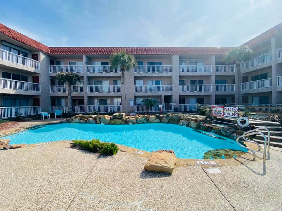 A lagoon-style swimming pool with clear blue water is surrounded by a textured concrete deck and rock features. The three-story building frames the pool area, with balconies on each floor offering access. Palm trees add a touch of greenery to the inviting outdoor space.