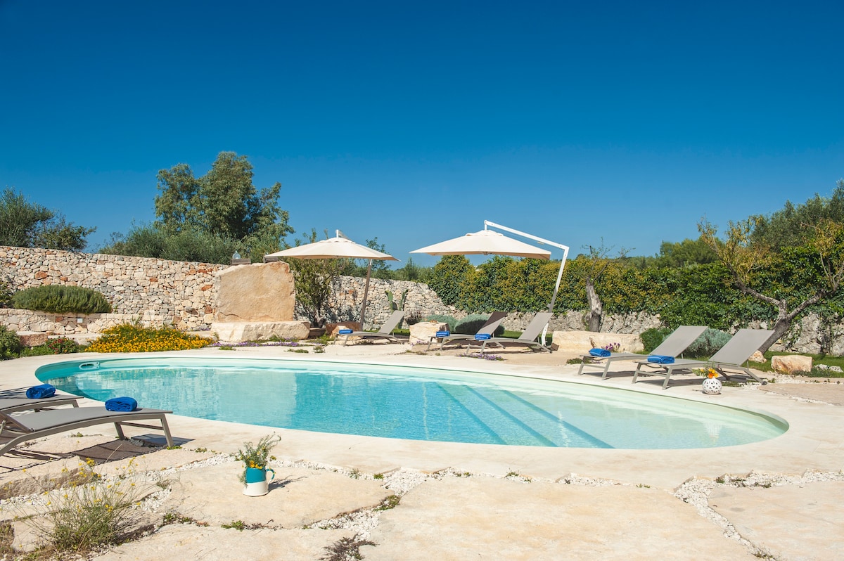 A spacious outdoor pool area features a tranquil swimming pool surrounded by sun loungers under large umbrellas. Lush greenery and flowering plants provide a natural backdrop, enhancing a serene environment for relaxation.