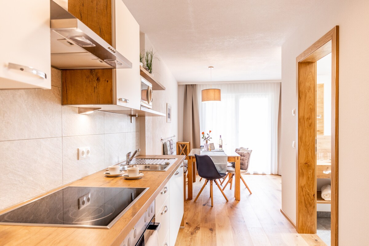 A modern kitchen area features sleek cabinetry and a spacious countertop with an electric stovetop. Natural light floods an adjacent dining space furnished with a wooden table and chairs. The open layout connects the kitchen to the living area, enhancing the overall sense of space.