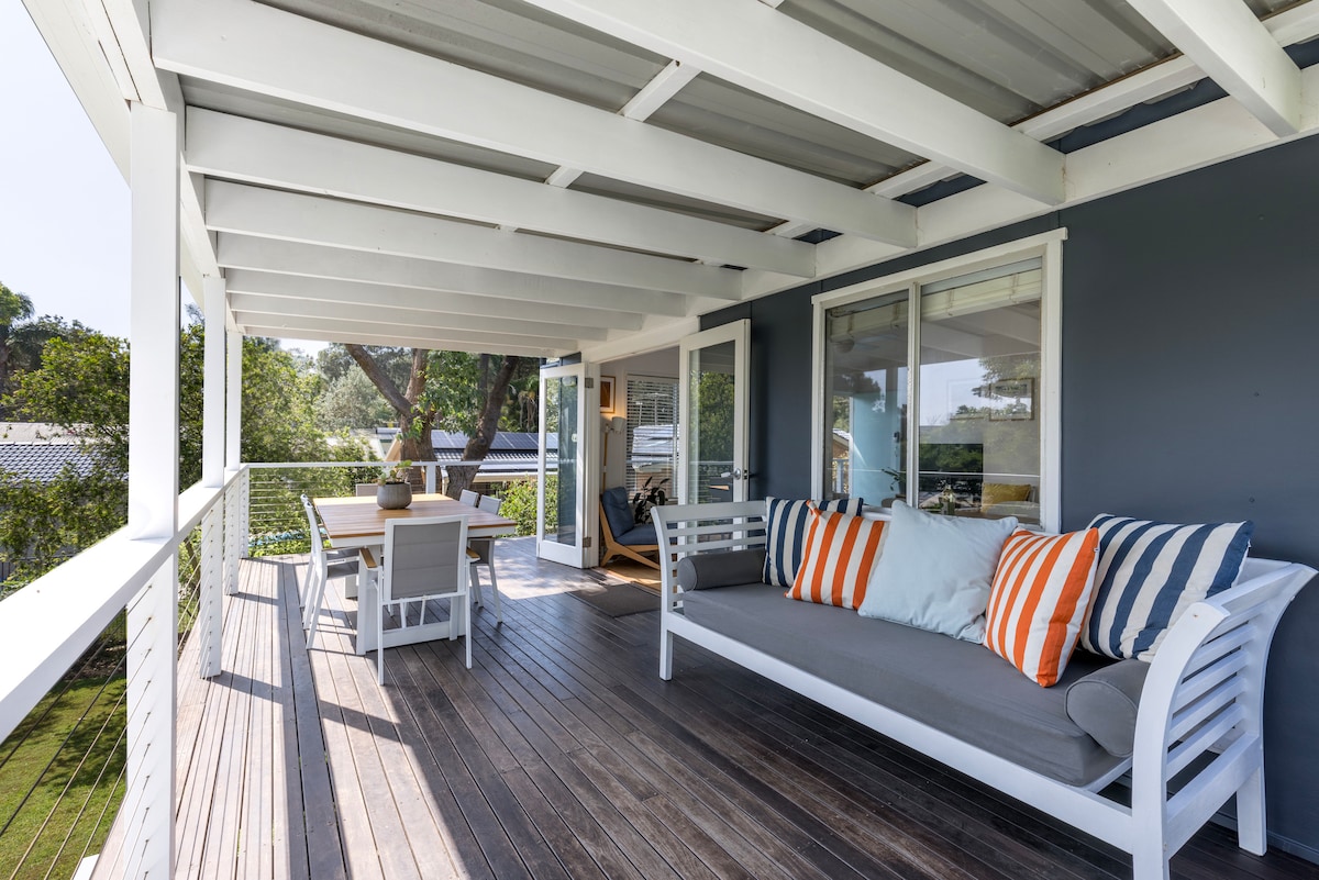 An outdoor deck is shown with a large daybed adorned with striped cushions, providing a comfortable spot to relax. A dining table set for six is positioned nearby. The space is surrounded by trees, enhancing the privacy and tranquility of this outdoor living area.