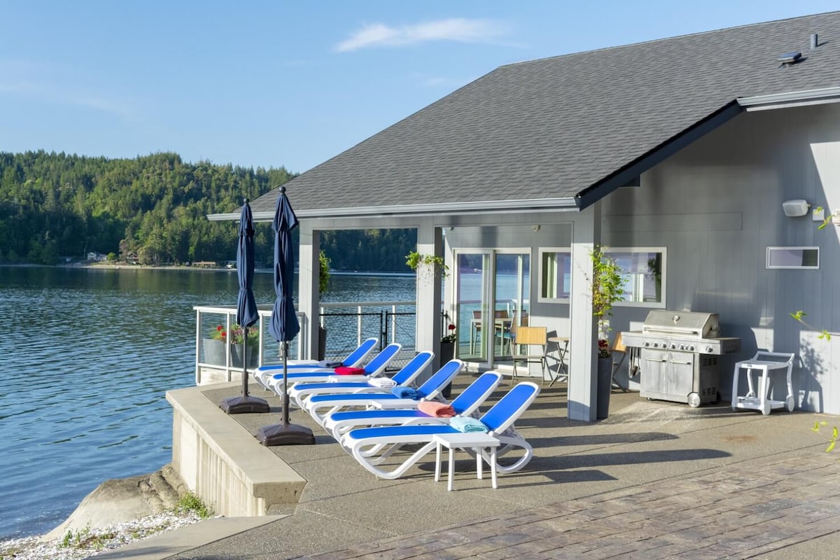 An outdoor area features several lounge chairs arranged around a patio overlooking the water. A gas barbecue grill is positioned nearby, and large patio umbrellas provide shade. The surroundings include a backdrop of trees and calm waters of Hood Canal.