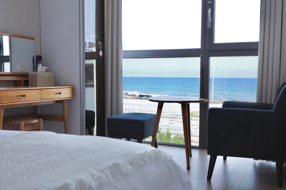 A spacious bedroom features a wooden vanity and a stylish armchair, complemented by a small round table. Large windows provide unobstructed views of the ocean and coastline, allowing natural light to brighten the room.