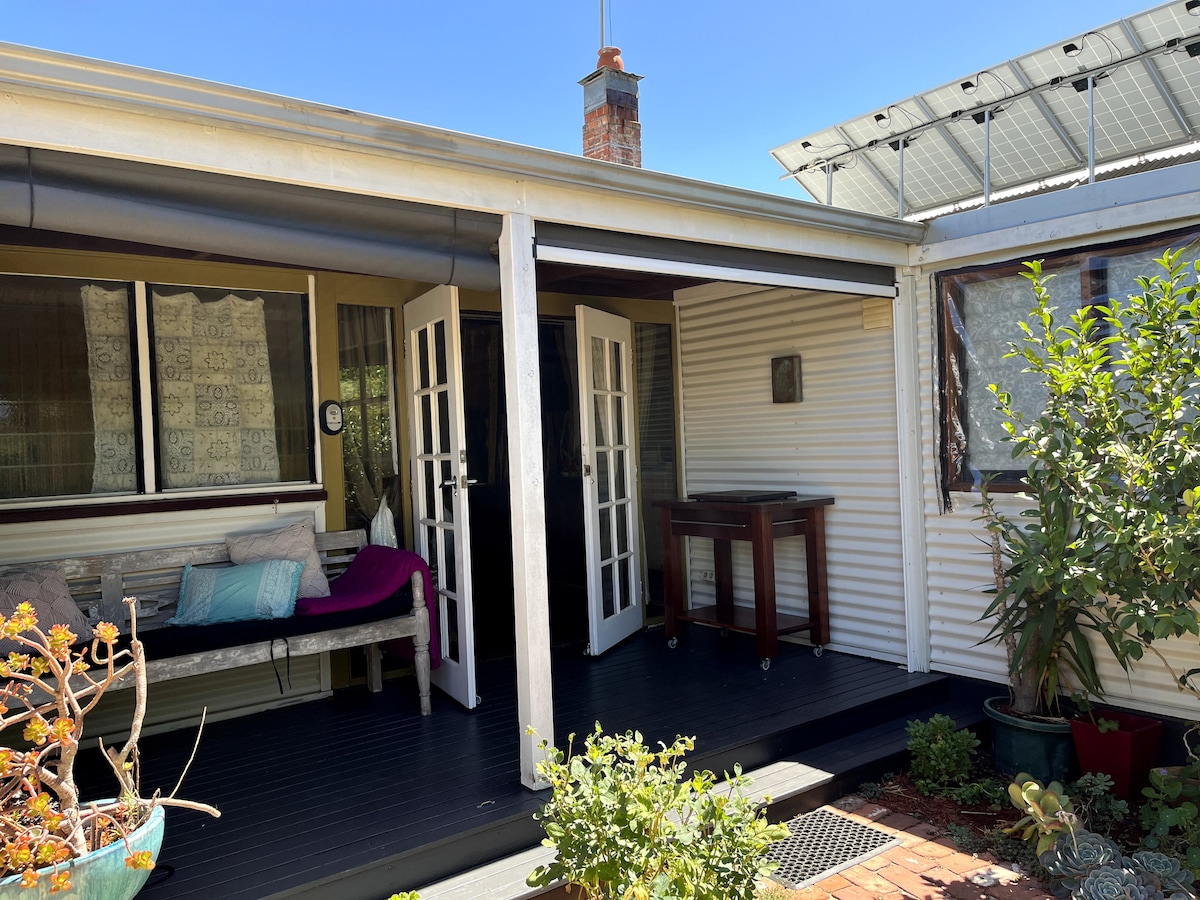The exterior features a covered entrance with two sets of French doors leading into the property. A bench sits to one side, complemented by potted plants and a small table. Sunlight illuminates the area, enhancing the inviting atmosphere.