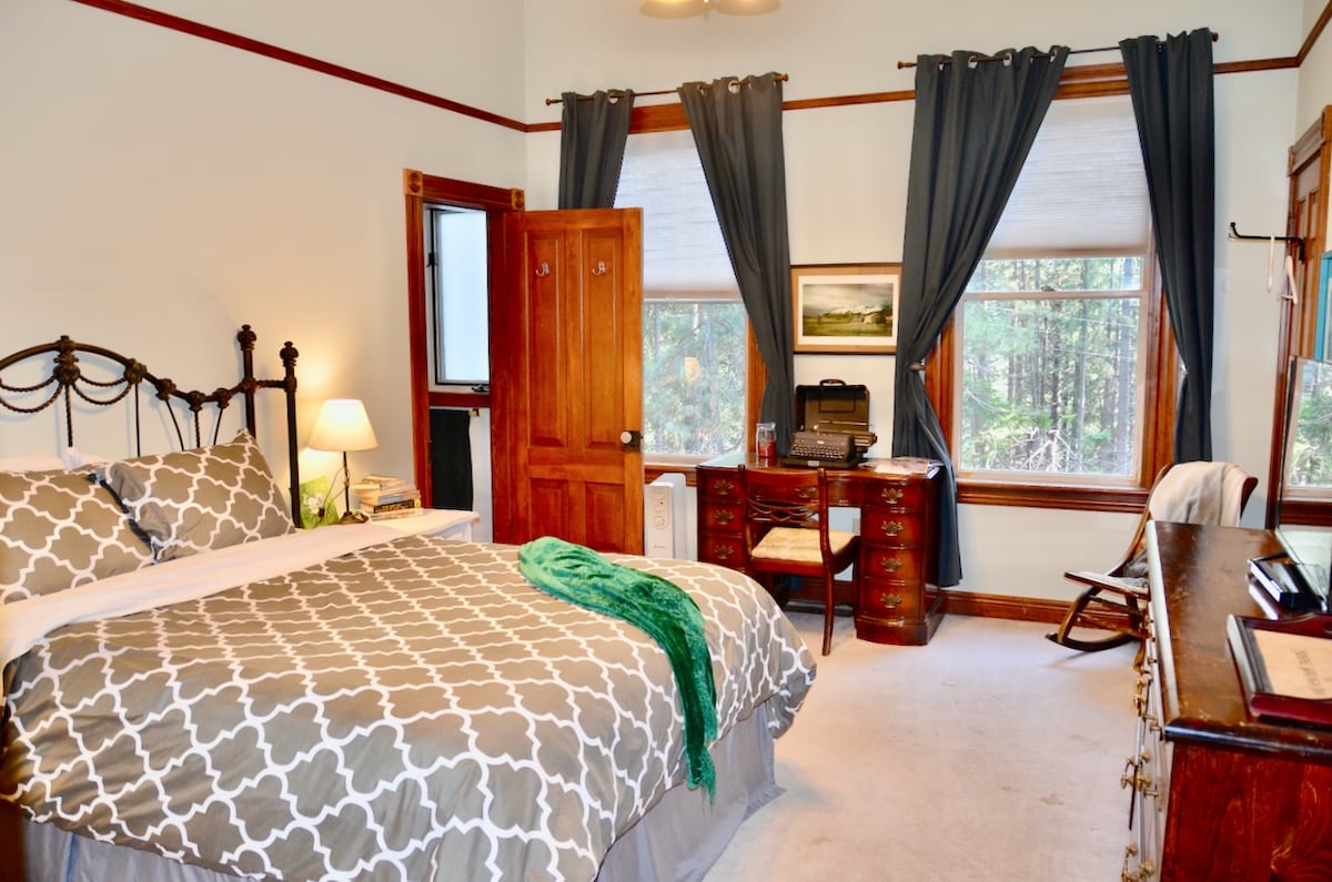 A well-appointed room features a spacious bed with patterned bedding. A wooden desk with a chair is positioned against a window, and two curtains frame the windows, allowing natural light. Decorative elements include a lamp and a vintage dresser, adding character to the space.