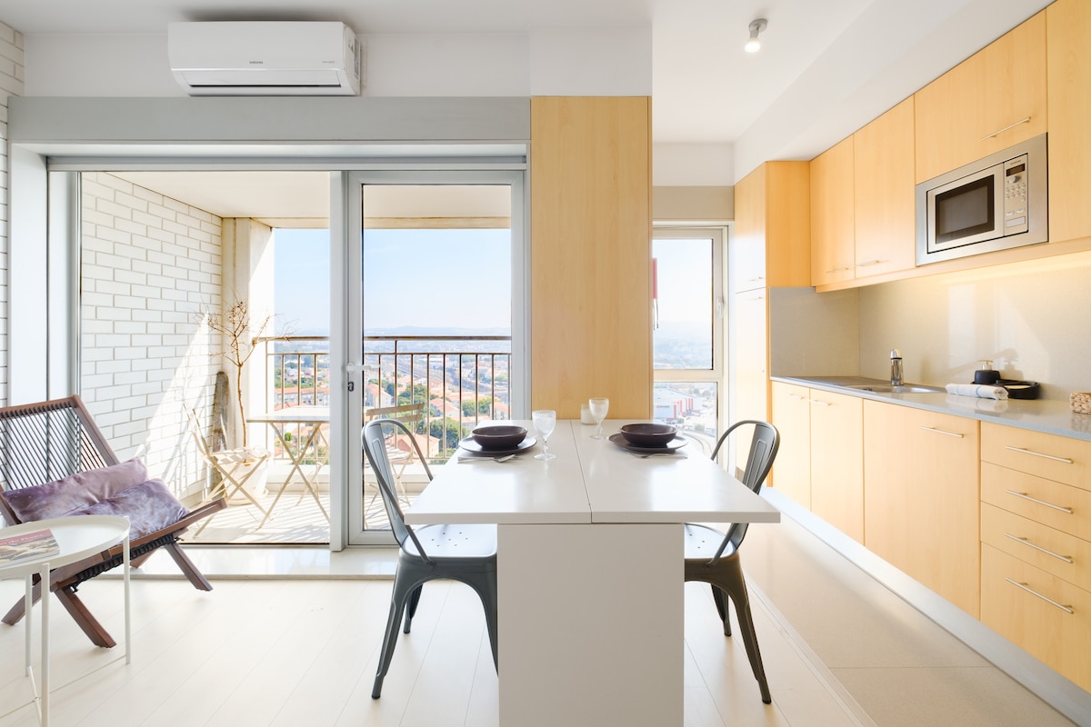 A bright and open kitchen area with light wooden cabinetry is complemented by a dining table set for two. Floor-to-ceiling glass doors lead to a balcony, offering picturesque views of the outdoors. A comfortable chair sits beside the table, enhancing the inviting space.