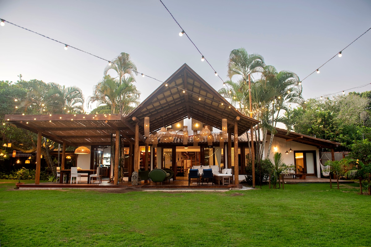 The spacious exterior of the property is framed by lush greenery and palm trees, with a striking architectural design featuring a large wooden structure. Soft outdoor lights are seen hanging above, enhancing the inviting appeal of the gathering space that opens to a well-maintained lawn.