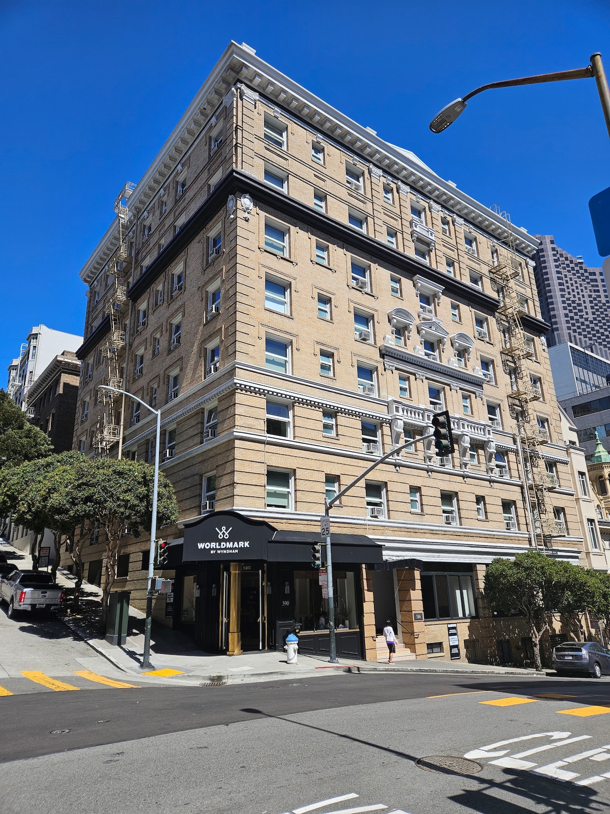 The exterior of the Worldmark San Francisco is captured from street level, showcasing a historic building with a striking facade and detailed architecture. The structure is framed by trees and sidewalks, with a bright blue sky above and a glimpse of the bustling streets.