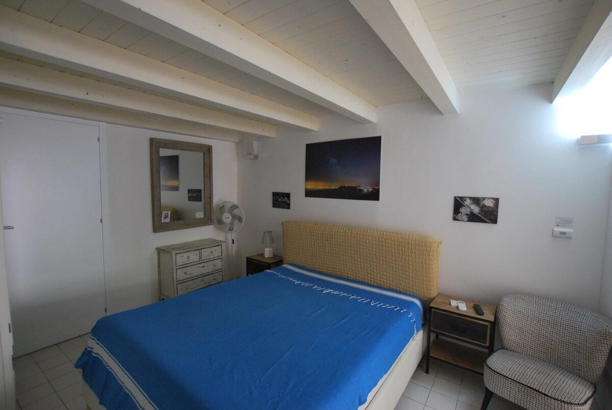 The sleeping area features a spacious bed with a blue blanket, flanked by wooden bedside tables and a patterned armchair. A dresser is positioned against the wall, complemented by a mirror above it. Natural light enters from a window, enhancing the clean and modern design.