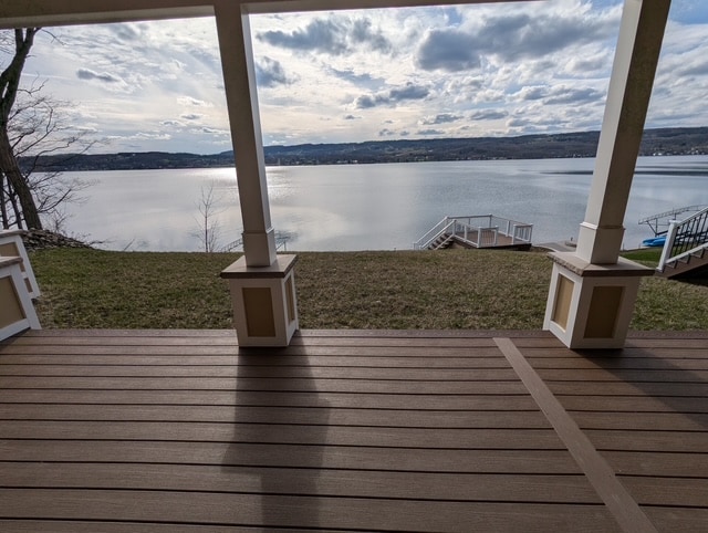 A private deck offers expansive views of the calm lake, framed by the supporting pillars of the structure. The serene water reflects the sky's soft clouds, creating a peaceful atmosphere. A grassy area extends from the deck toward the lake, leading to stairs that descend to the water.