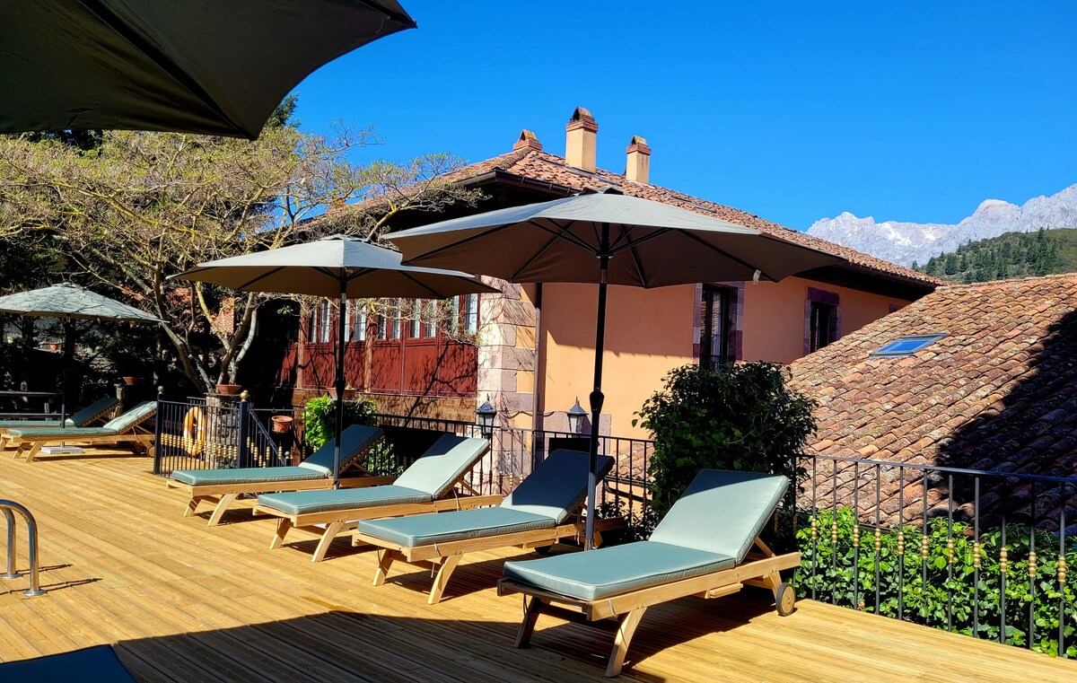 A sunlit terrace features several loungers positioned under large umbrellas. The wooden decking surrounds a swimming pool, while traditional buildings with red and ochre facades are visible in the background, complemented by lush greenery and distant mountain peaks.