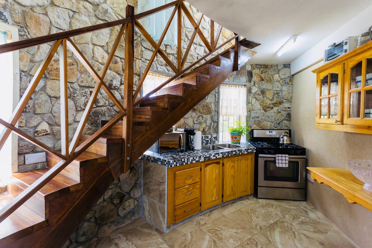 The kitchen area features wooden cabinetry with stone accents, complementing the rustic charm of the setting. A staircase with wooden railing leads to the upper level, while natural light enters through the nearby window and enhances the room's warm tones.