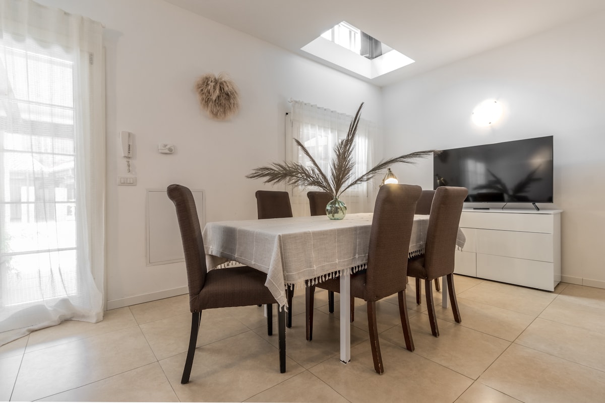 A dining area is presented, featuring a rectangular table set with six upholstered chairs. Natural light enters through a skylight above, illuminating the space. A flat-screen television is mounted on the wall, and decorative elements, such as a vase with pampas grass, enhance the inviting atmosphere.