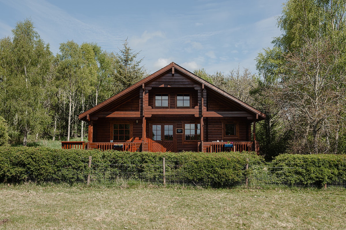 A wooden log cabin stands amidst serene greenery, featuring a large front veranda with outdoor seating. The structure is flanked by trees and a well-maintained hedge, with large windows allowing natural light to enter the inviting interior.