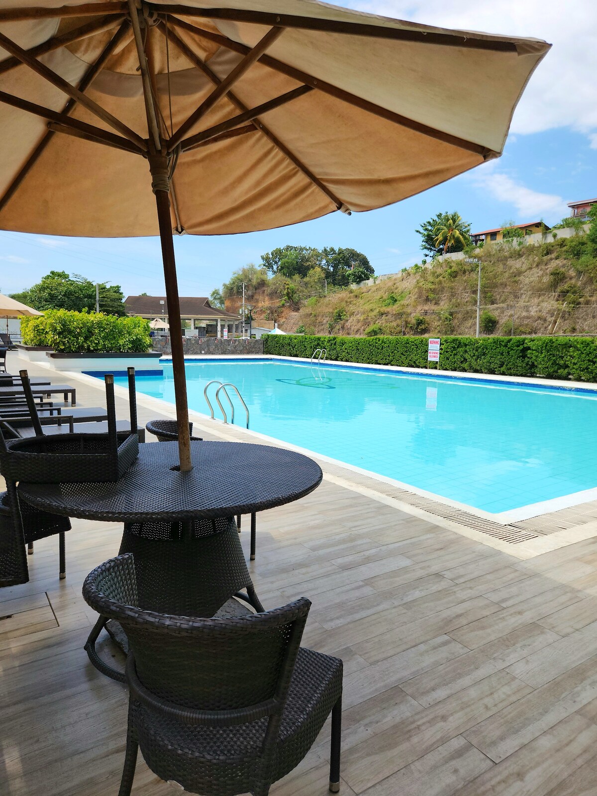 A large swimming pool is bordered by lush greenery and lounge chairs under a shaded parasol. A stone pathway leads to the pool area, creating a serene atmosphere. The setting invites relaxation and enjoyment of the outdoor space.