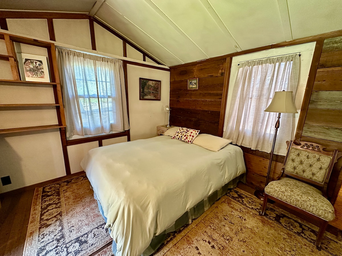 An open-space room features a spacious bed with soft linens and a decorative pillow. Natural light filters through two windows, complemented by sheer curtains. A wooden shelf stands against one wall, and a comfortable chair with a patterned upholstery is positioned nearby, enhancing the cozy atmosphere.