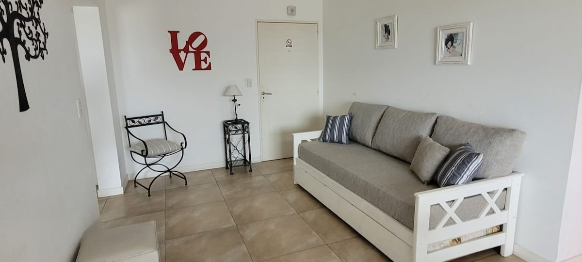 A light-filled living area features a gray sofa adorned with striped cushions, complemented by a decorative chair. A black side table and wall art add character, while a 'LOVE' sign brings a touch of warmth to the ambiance.