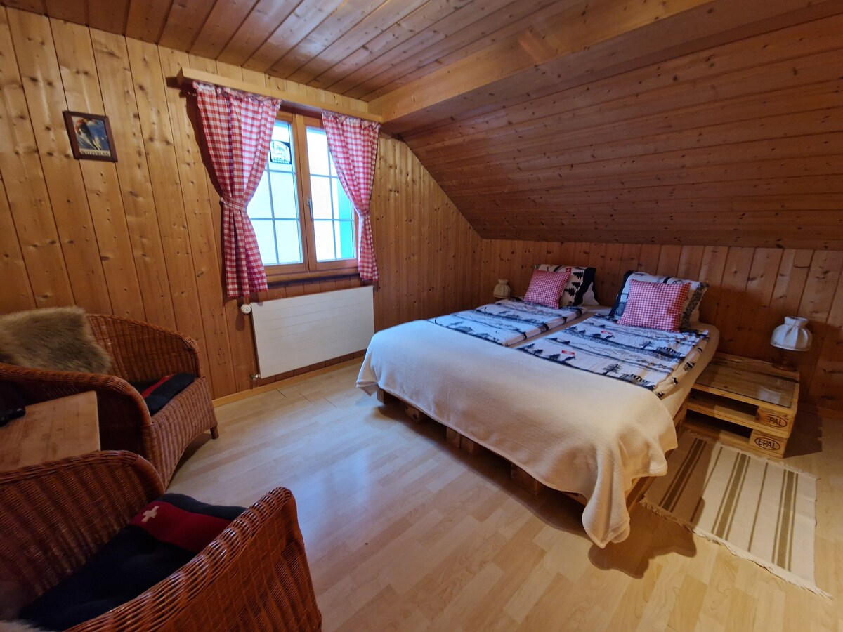 A cozy double room features two single beds with patterned bedding. A pair of wicker chairs are positioned near a small table. Wooden walls and ceiling create a warm atmosphere, while natural light enters through two windows adorned with checkered curtains.