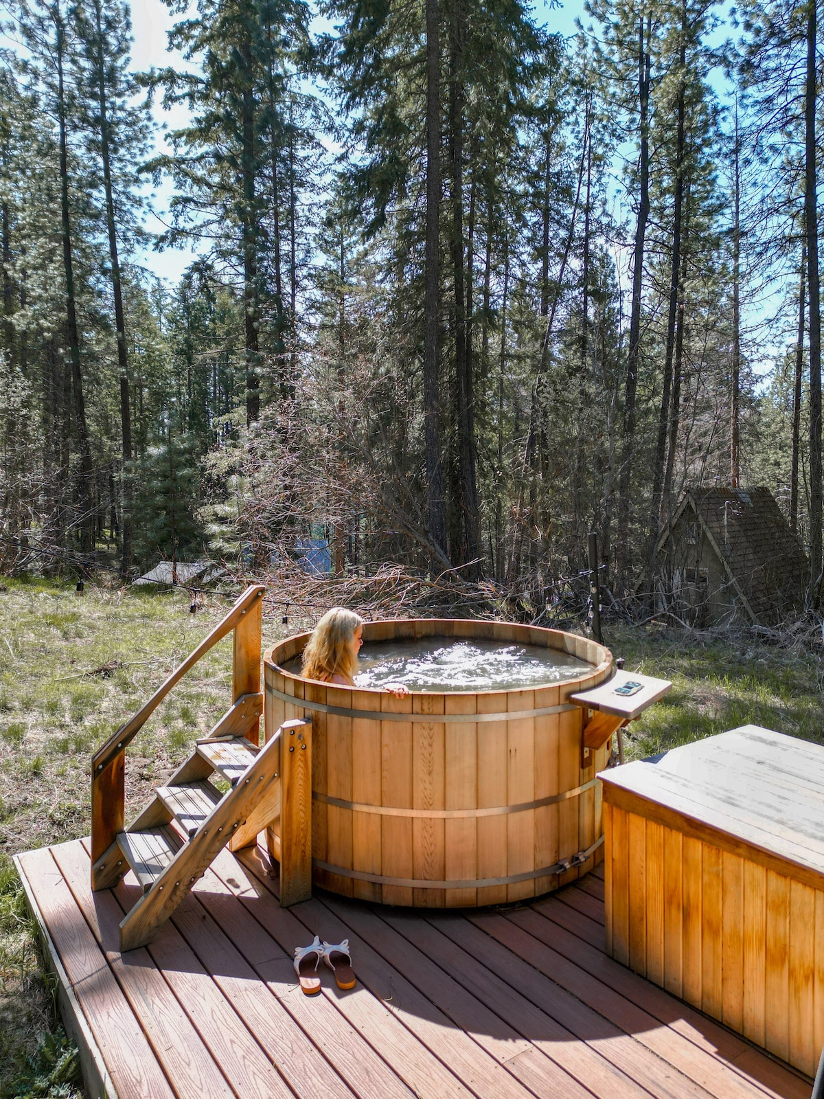 A cedar hot tub is set on a wooden deck, surrounded by tall trees and foliage. Steps lead up to the hot tub, which appears to be filled with water. A pair of shoes sits on the deck nearby, enhancing the outdoor, relaxed atmosphere.
