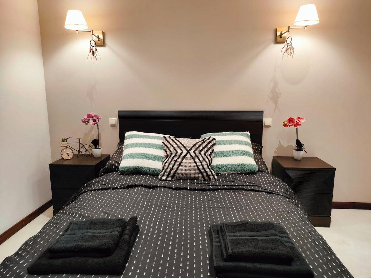 A comfortable sleeping area features a spacious bed with striped pillows and a dark bedspread. Two nightstands are positioned on either side of the bed, adorned with decorative orchids. Soft lighting is provided by wall sconces, enhancing the serene atmosphere.
