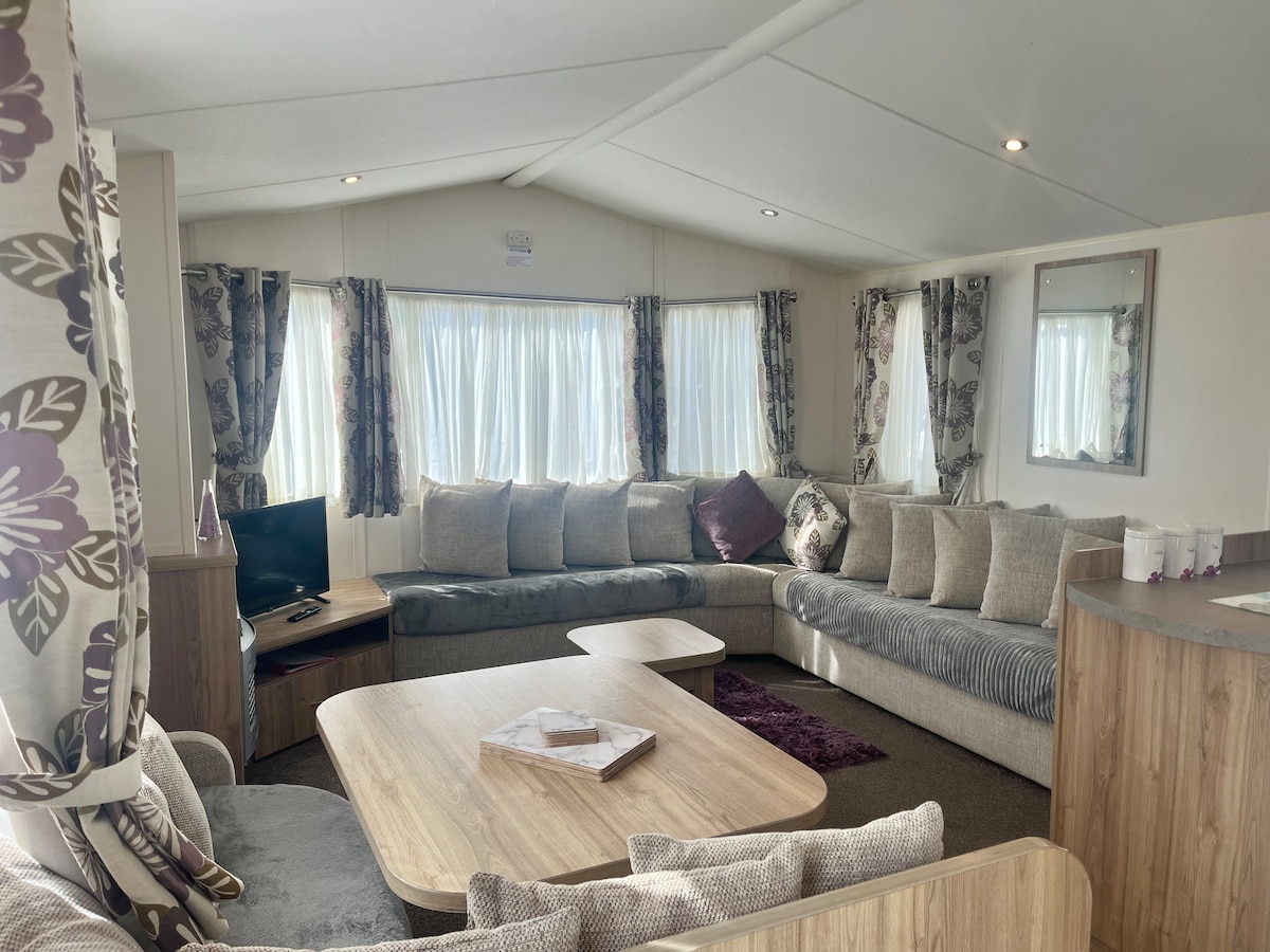 A spacious living area features a large, L-shaped sofa adorned with decorative cushions. A coffee table sits in the center, complemented by a TV in the corner. Natural light floods through large windows, framed by stylish curtains, creating a bright and welcoming environment.