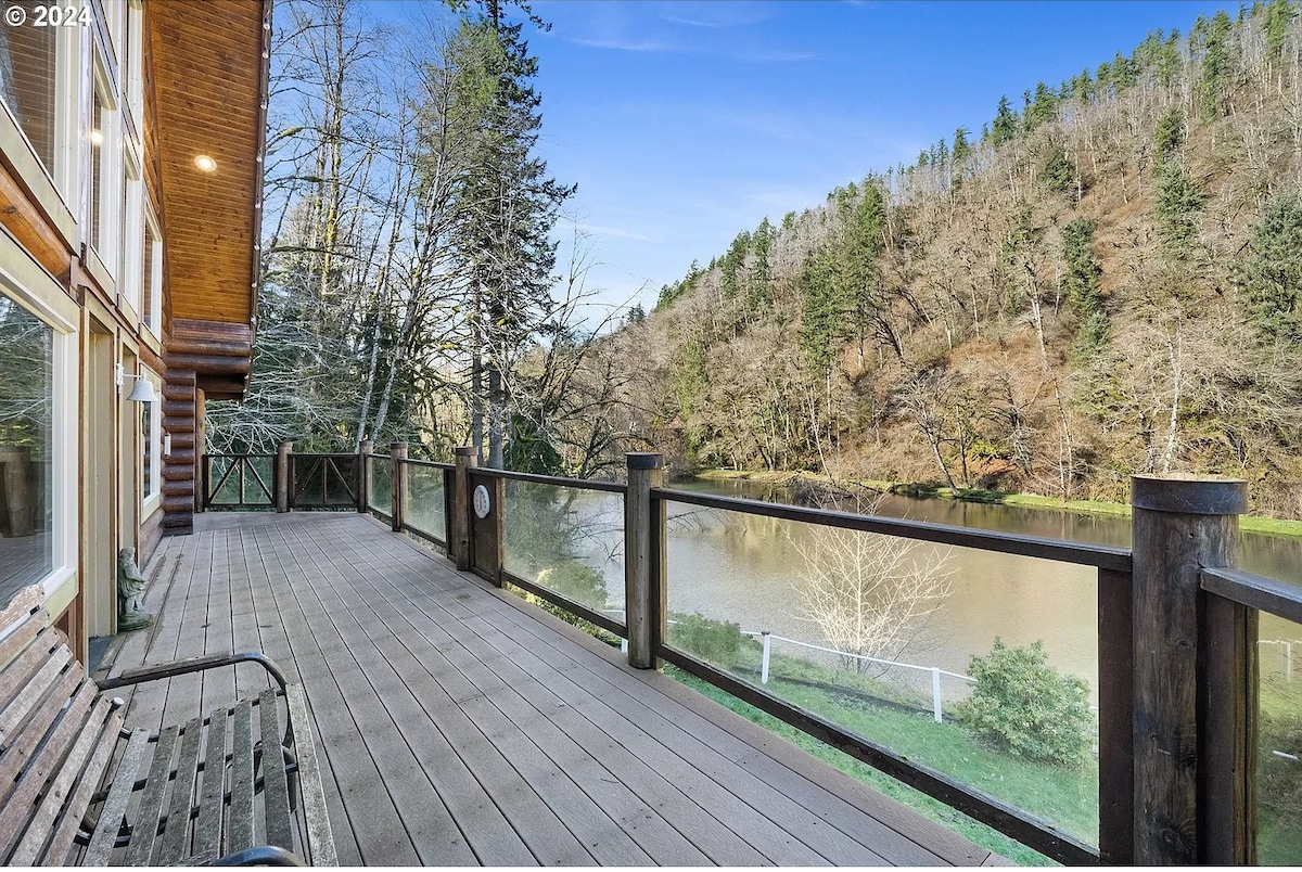 A spacious wooden deck is featured, overlooking a serene body of water surrounded by a forested hillside. The railing provides a clear view of the landscape, where trees create a natural frame for the tranquil scene.