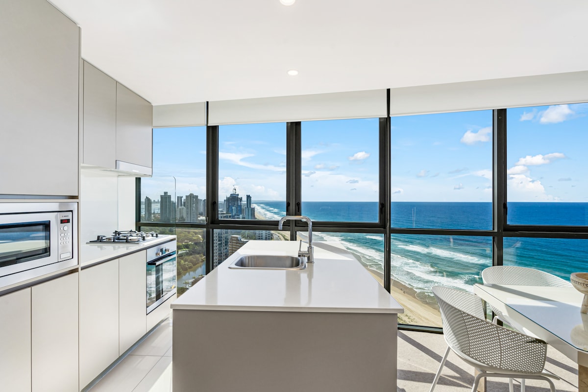 A modern kitchen is highlighted by floor-to-ceiling windows offering panoramic views of the Pacific Ocean and the Northern Gold Coast skyline. The space includes sleek cabinetry, a gas stove, an oven, and a small dining area with a round table, allowing for effortless entertaining.
