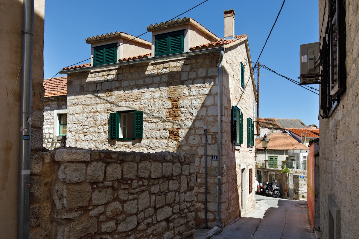 A charming stone house stands at the end of a narrow alley, featuring green wooden shutters on large windows. The building showcases a blend of traditional architecture with a weathered exterior, complemented by the clear blue sky above.