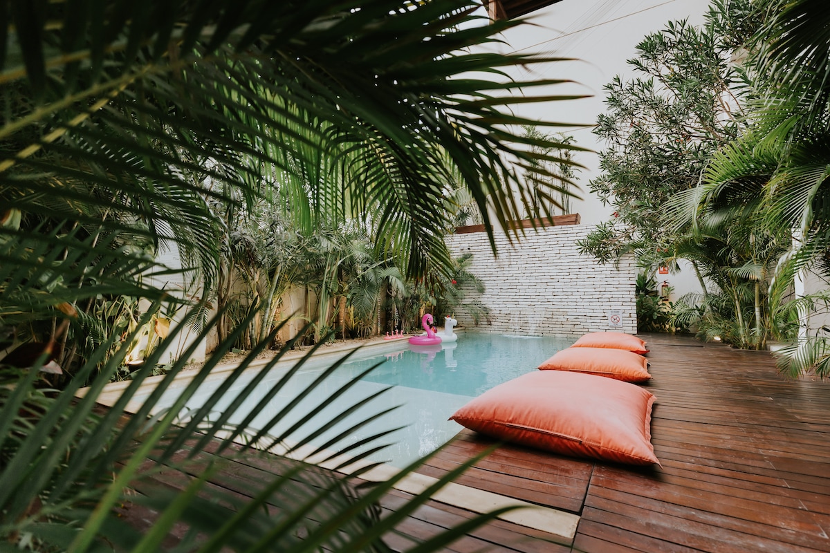 A private pool surrounded by lush tropical plants is featured, with vibrant orange cushions placed on a wooden deck. The tranquil water reflects the greenery, creating a serene atmosphere. A gentle splash from a guest enjoying the pool can be seen, enhancing the inviting space.
