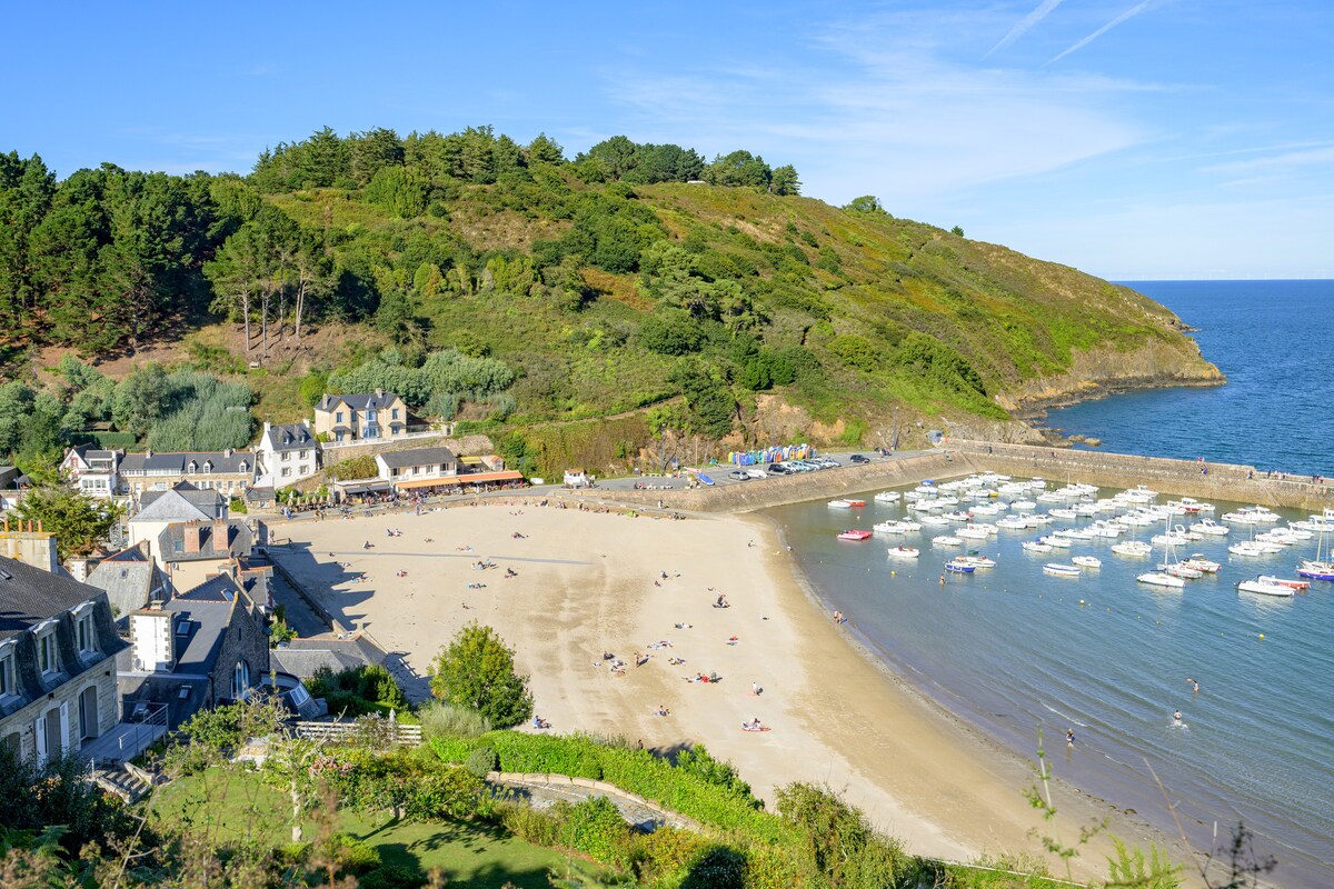 A scenic view captures the soft sandy beach alongside calm waters, dotted with small boats. Lush green hills rise in the background, and quaint buildings are positioned along the shoreline, providing a peaceful coastal atmosphere.