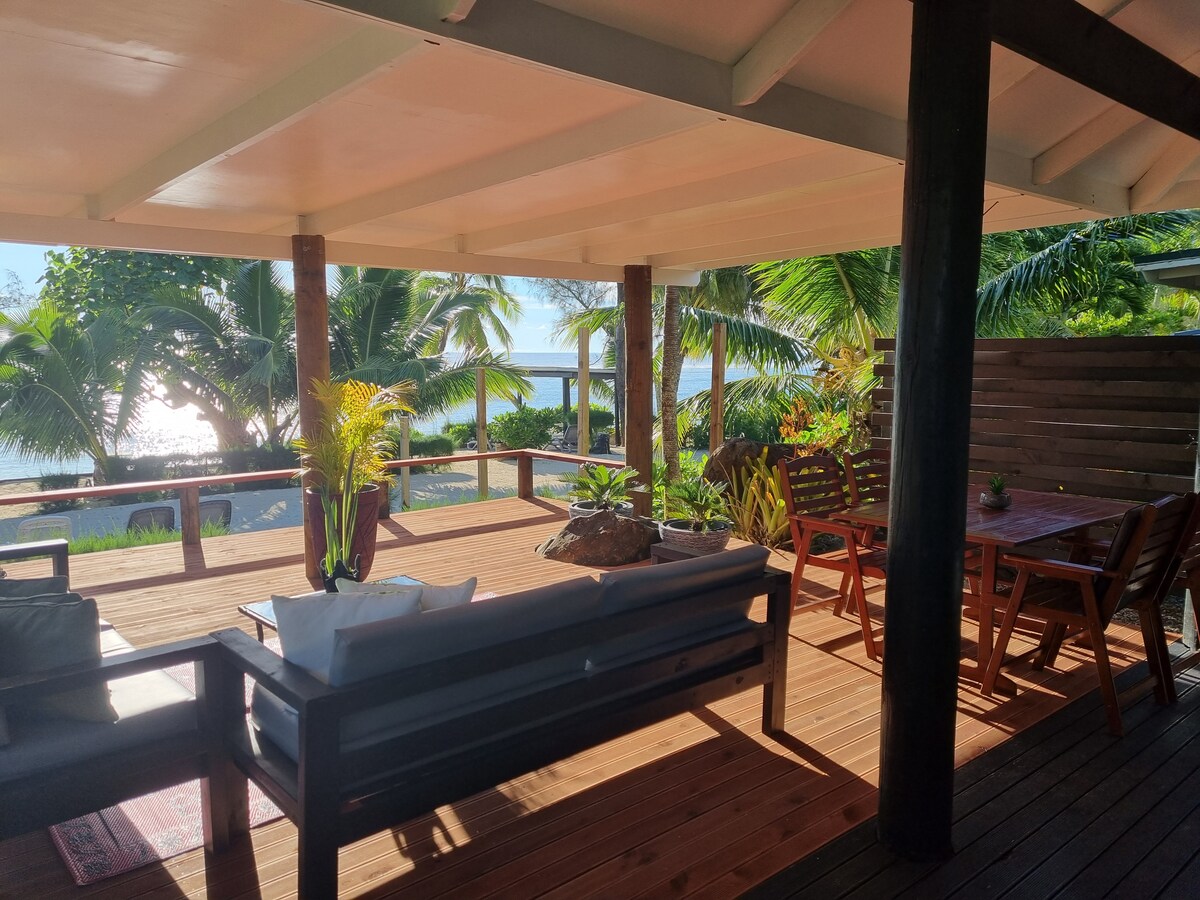 A spacious decking area features comfortable seating and a dining table, surrounded by tropical greenery and palm trees. The serene ocean view is visible in the background, enhancing the outdoor living space perfect for relaxing or dining.
