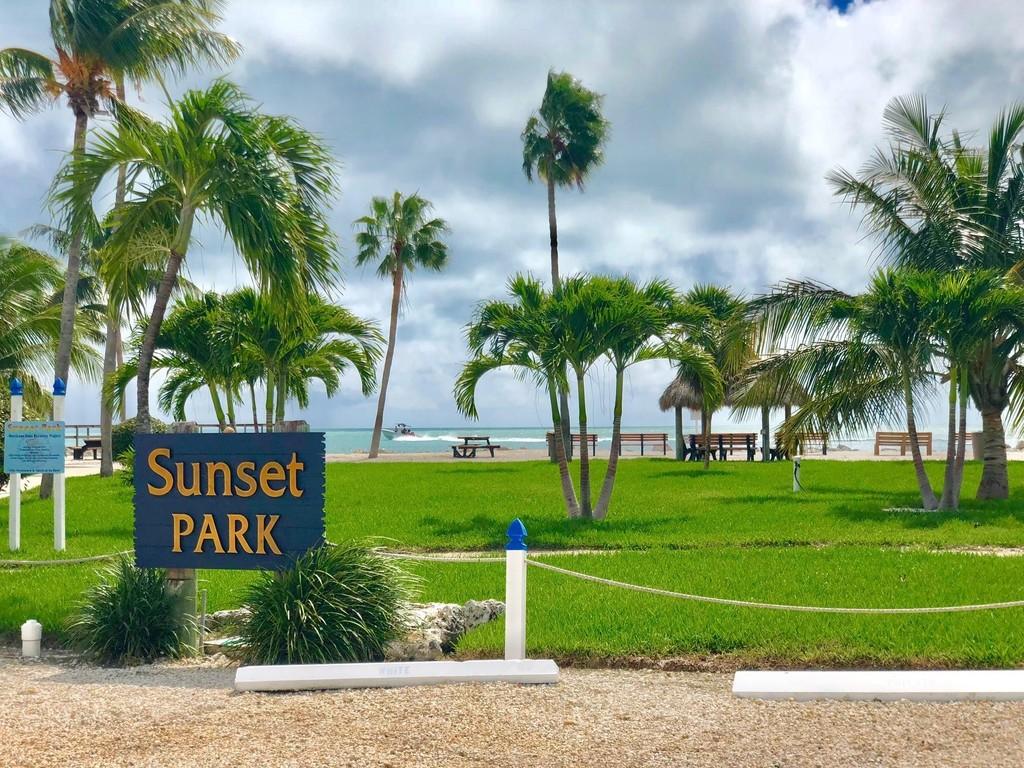 A grassy area is surrounded by palm trees, with a sign displaying 'Sunset Park' prominently in front. Wooden picnic tables are positioned in the park, with the ocean visible in the background under a partly cloudy sky.