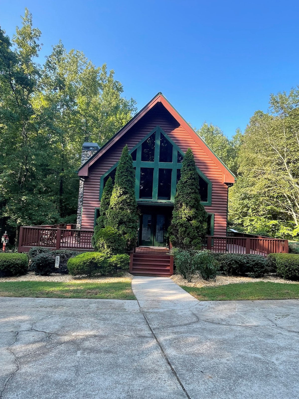 The exterior of the cabin features a triangular roof and large, green-tinted windows that allow natural light to enter. Surrounding the cabin are neatly trimmed shrubs and mature trees, providing a sense of privacy. A spacious wooden deck enhances the welcoming entrance.