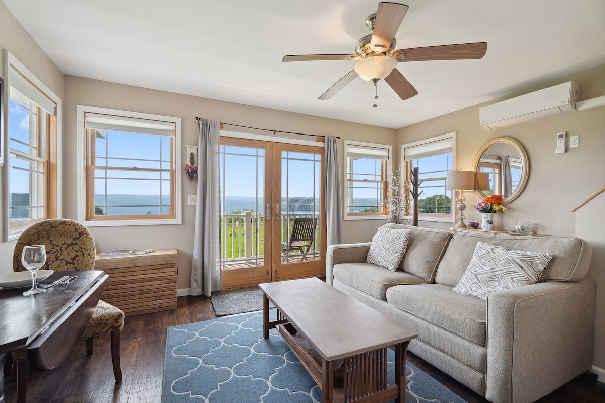 A living area is presented with a comfortable sofa and a coffee table, overlooking expansive views of Seneca Lake through large windows. French doors lead to an outdoor deck, while decorative elements like a patterned rug and a ceiling fan add to the welcoming ambiance.