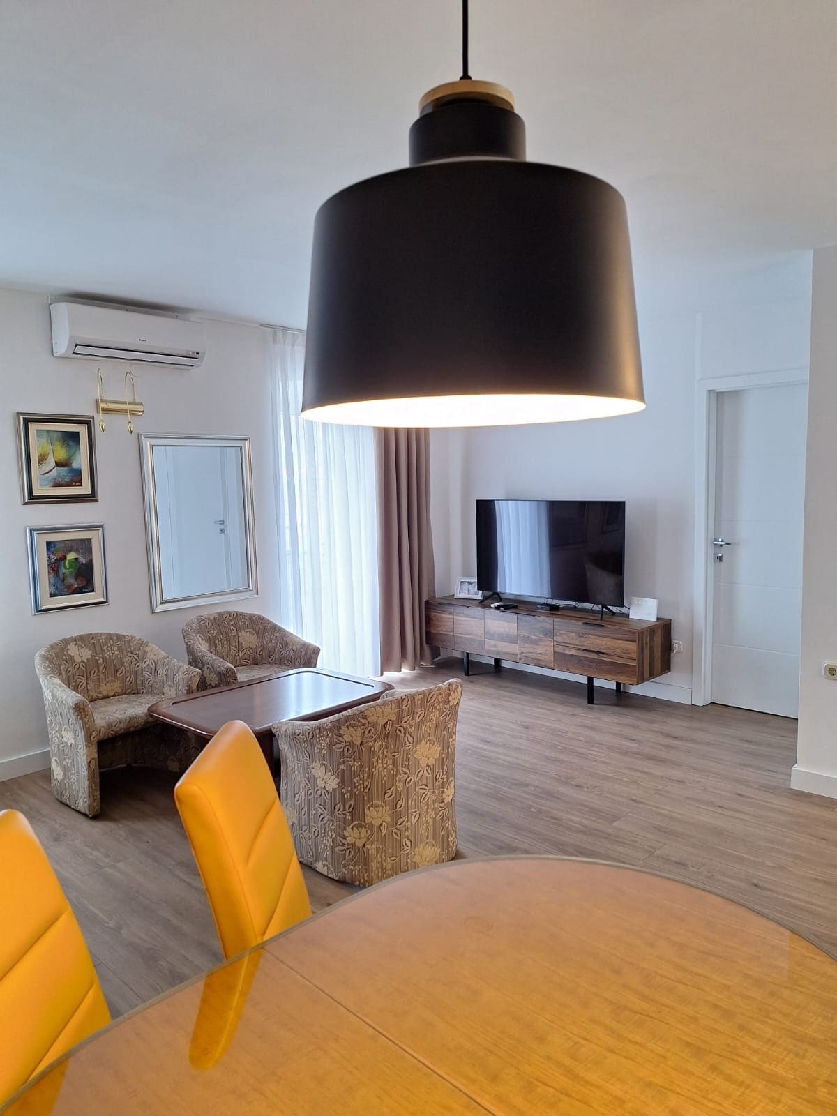 A well-lit living area is presented, featuring a modern television mounted on a wooden console. Two armchairs and a dining table are visible, with yellow chairs enhancing the space. Natural light filters in through sheer curtains, softly illuminating the room's neutral decor.