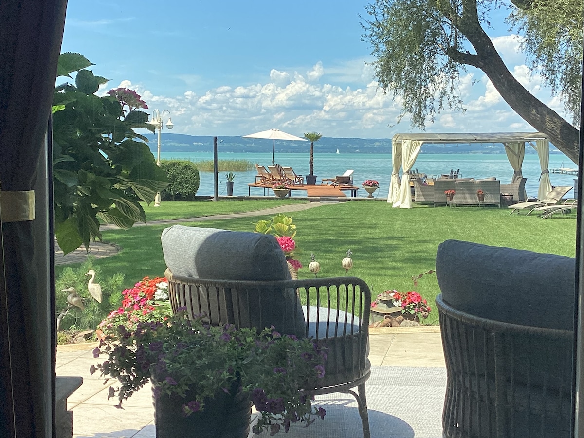 A serene outdoor space is presented with comfortable seating facing a vast lawn. Colorful flowers line the path to a private dock featuring sun loungers and an umbrella. The horizon reveals calm waters of the Balaton under a partly cloudy sky.