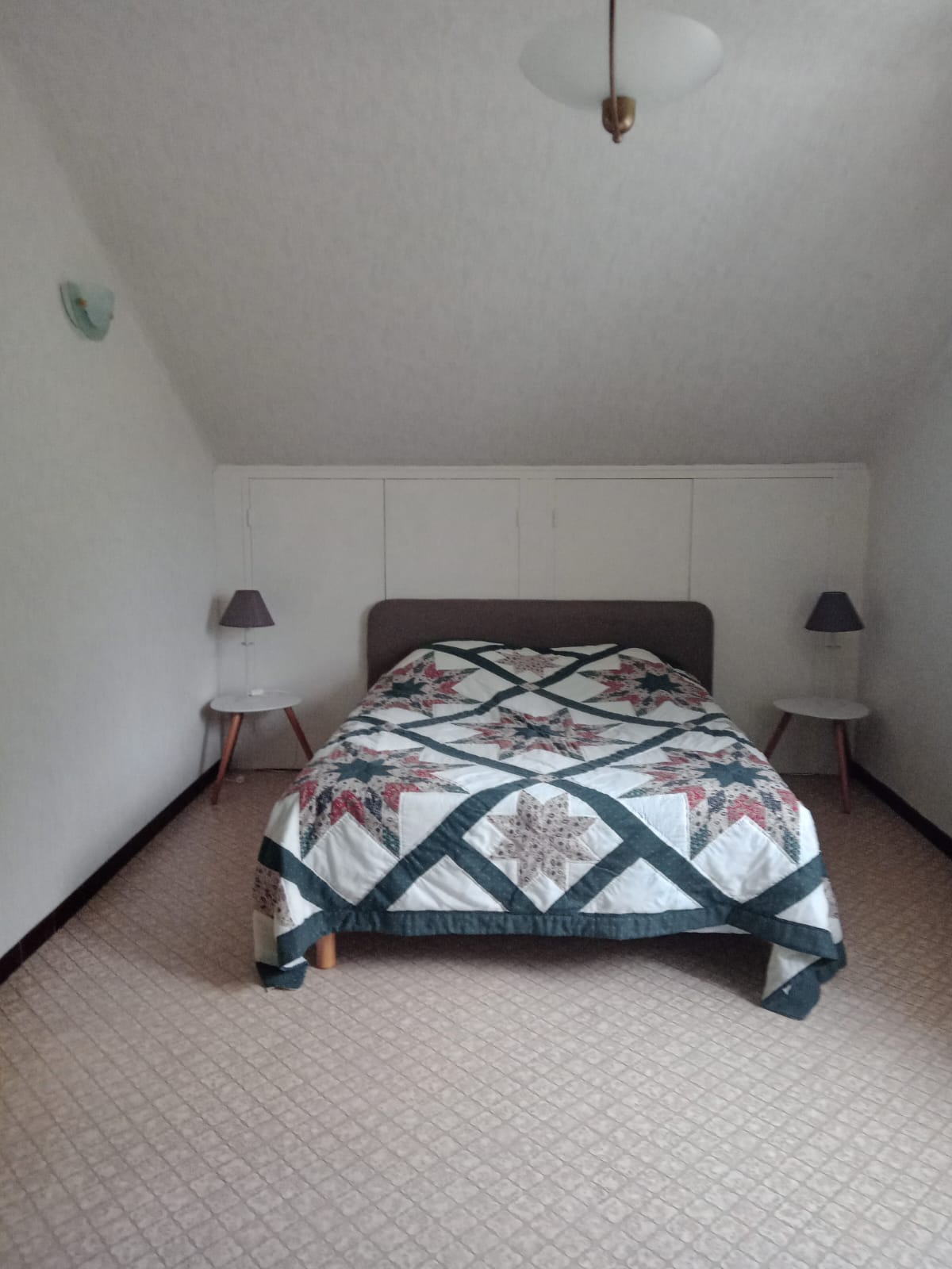 A cozy bedroom features a spacious bed covered by a patterned quilt. Two bedside tables with lamps are positioned on either side. The room showcases subtle wall decor and a carpeted floor, complemented by a gently sloping ceiling.