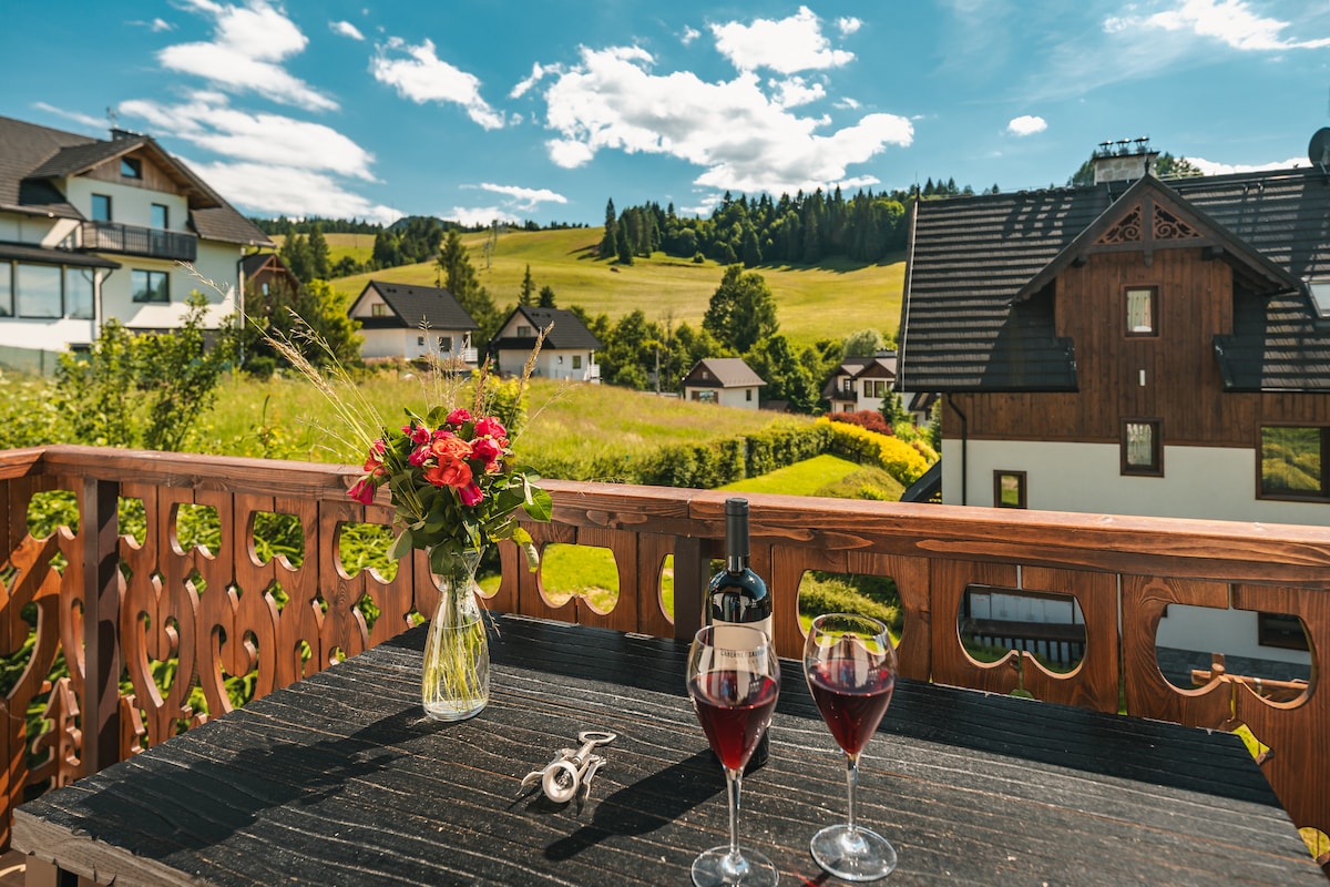 A balcony table is set with two glasses of red wine and a bottle, accompanied by a vase of fresh roses. Rolling green hills and quaint houses are visible in the background under a bright blue sky.