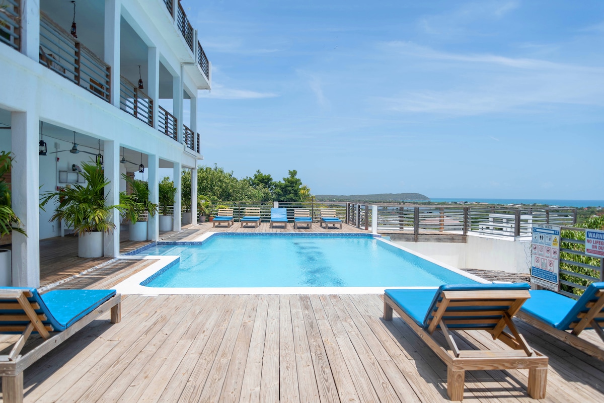 A tranquil infinity pool is set against a backdrop of lush greenery and ocean views. Sun loungers with blue cushions are arranged around the pool area, complemented by the wooden deck. The building features multiple levels, adorned with balconies that overlook the inviting outdoor space.